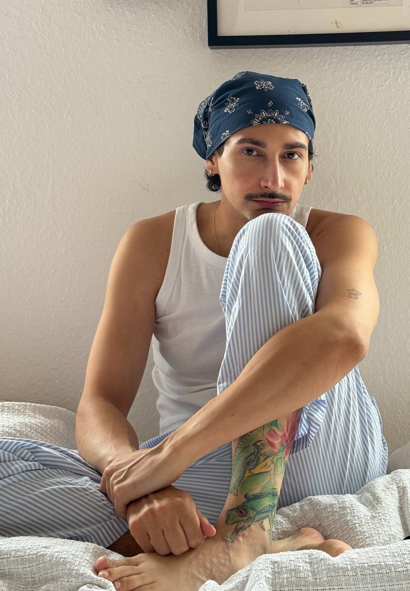 Junge Person sitzt auf dem Bett, trägt ein blaues Bandana, ein weißes Tanktop, gestreifte Hosen und zeigt einfarbiges Tattoo auf dem Bein.
