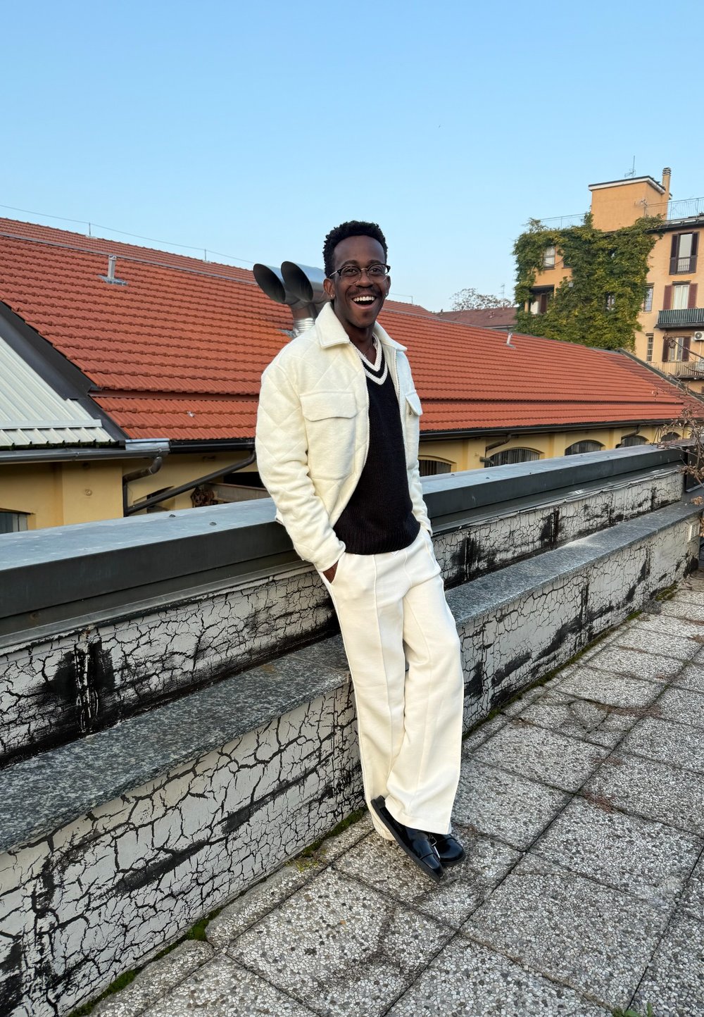 Giacca di pile bianca, maglione nero a scollo a V, pantaloni bianchi su misura e scarpe nere. Una persona sorridente posa contro un muro testurizzato e un tetto.