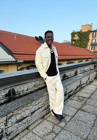 Giacca di pile bianca, maglione nero a scollo a V, pantaloni bianchi su misura e scarpe nere. Una persona sorridente posa contro un muro testurizzato e un tetto.
