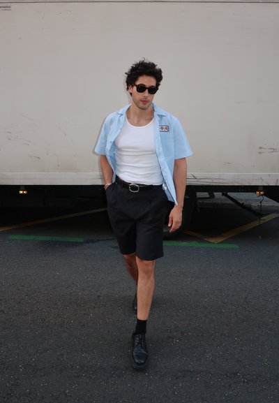 Camisa de manga corta azul claro, camiseta blanca sin mangas, pantalones cortos negros y zapatos negros. Con las manos en los bolsillos, de pie sobre una superficie pavimentada.