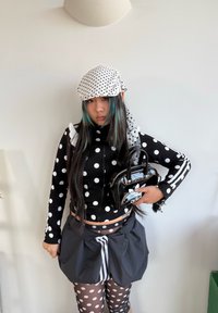 Musta pilkullinen vetoketjutakki, jossa on röyhelökaulus, mustavalkoiset pilkulliset leggingsit, ylisuurikokoiset mustat shortsit ja kiiltävä musta käsilaukku.