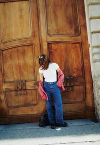 T-shirt bianca sotto una camicia rossa a righe aperta, jeans blu a vita alta, scarpe nere, sullo sfondo di una grande porta di legno.