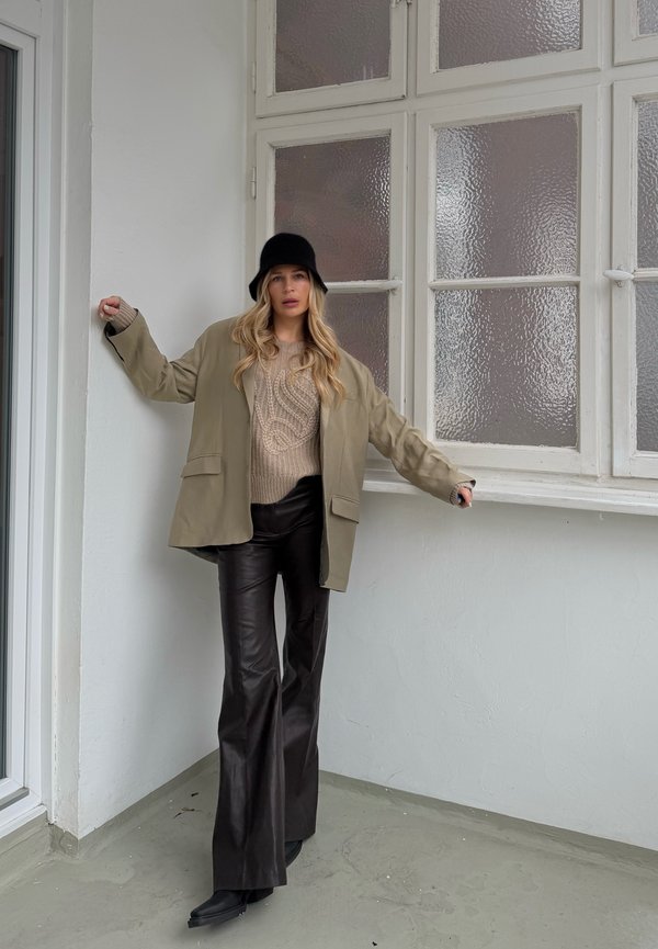 Donna con cappello a secchiello nero, blazer beige, maglione a maglia marrone chiaro e pantaloni in pelle nera, in piedi all'interno vicino a finestre con vetro smerigliato.