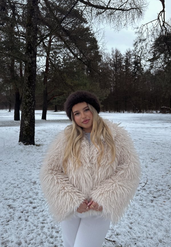 Młoda kobieta w puszystym, beżowym płaszczu i ciemnej futrzanej czapce stoi na śnieżnej ziemi z drzewami w zimowym lesie.