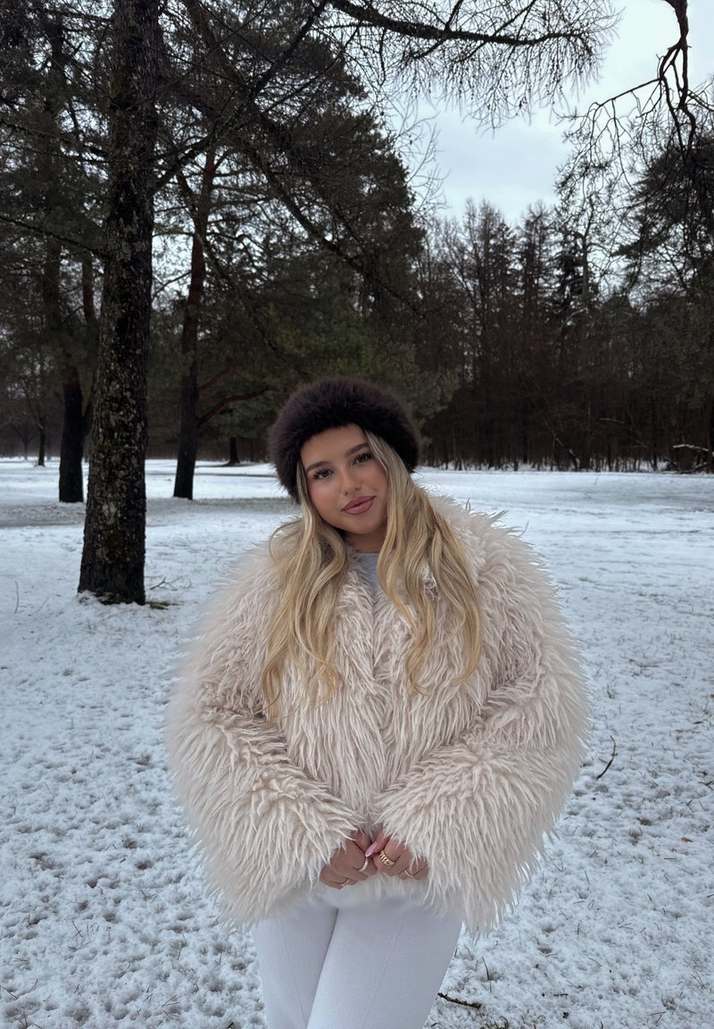 Jeune femme en manteau beige duveteux et chapeau en fourrure foncée, debout sur un sol enneigé avec des arbres dans un cadre de forêt hivernale.