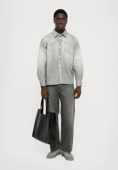 Chaqueta de denim gris con cierre de botones en la parte delantera, combinada con jeans grises. El modelo sostiene un bolso tote de cuero negro con dos asas.