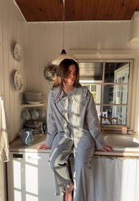 Outfit a righe in grigio chiaro e scuro, con colletto volantino e vestibilità ampia. Lo scenario include elementi di cucina rustica e luce solare.