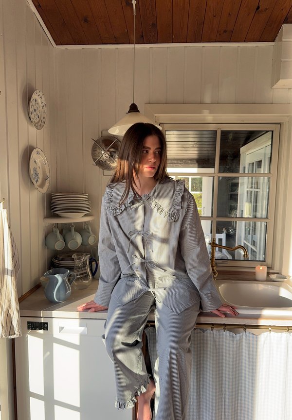 Lined outfit in light and dark grey, featuring a frilled collar and a relaxed fit. The setting includes rustic kitchen elements and sunlight.
