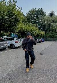 Čierny rebrovaný sveter, tmavé džínsy a béžové topánky. Čierny šiltovka a taška cez telo. Spevnená parkovacia plocha s autami a zeleňou.