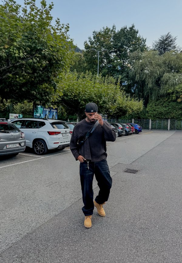 Čierny rebrovaný sveter, tmavé džínsy a béžové topánky. Čierny šiltovka a taška cez telo. Spevnená parkovacia plocha s autami a zeleňou.