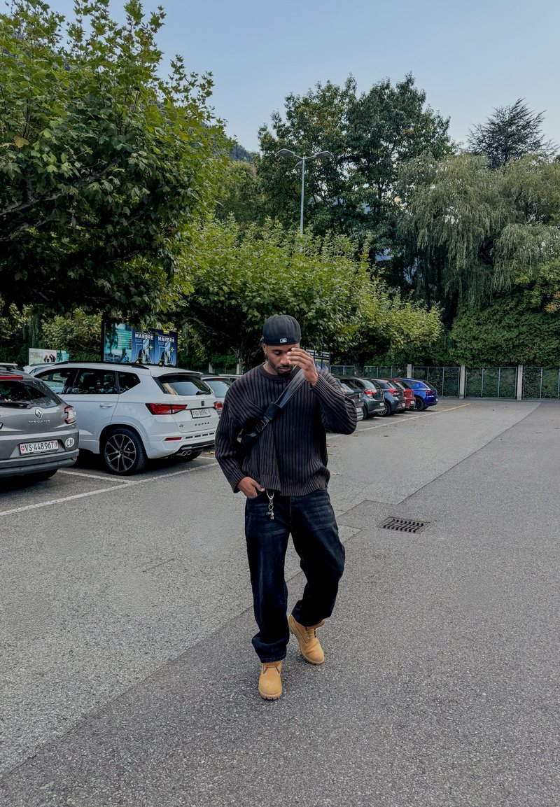 Suéter preto canelado, jeans de ganga escuros e botas castanhas. Boné preto e bolsa de ombro. Área de estacionamento pavimentada com carros e vegetação.