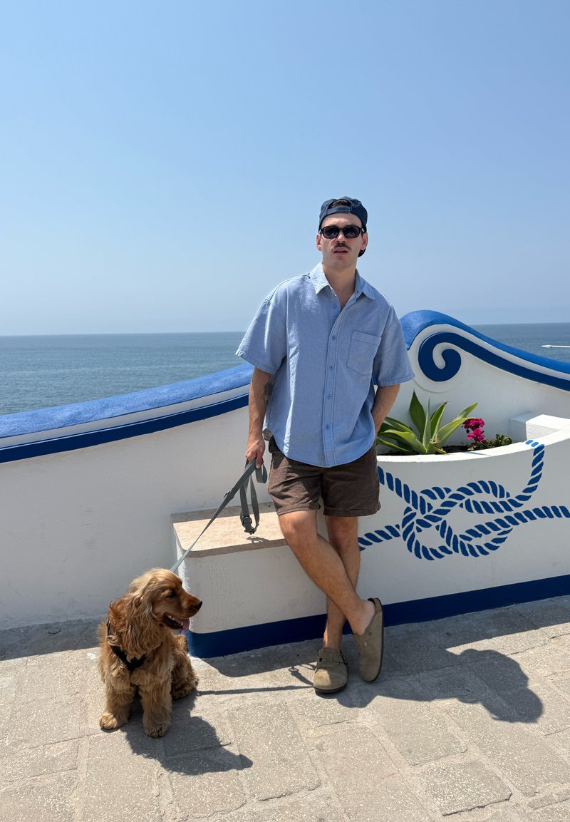 Homme en lunettes de soleil et casquette se tient près d'un mur de mer bleu et blanc, tenant la laisse d'un chien doré assis avec l'océan en arrière-plan.