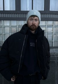 Veste matelassée noire avec un col montant, associée à un sweat-shirt bleu marine foncé avec un texte blanc. Bonnet gris clair. Fond neutre.