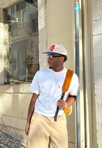 White short-sleeved T-shirt, beige shorts, light brown fuzzy backpack strap, clear glasses, gold necklace, standing near a textured wall.