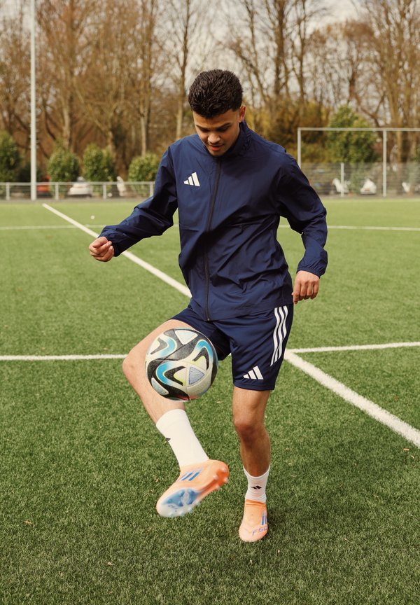 Mladý muž v námořnicky modrém sportovním oblečení žongluje fotbalovým míčem na zeleném pažitu s holými stromy a brankami v pozadí.