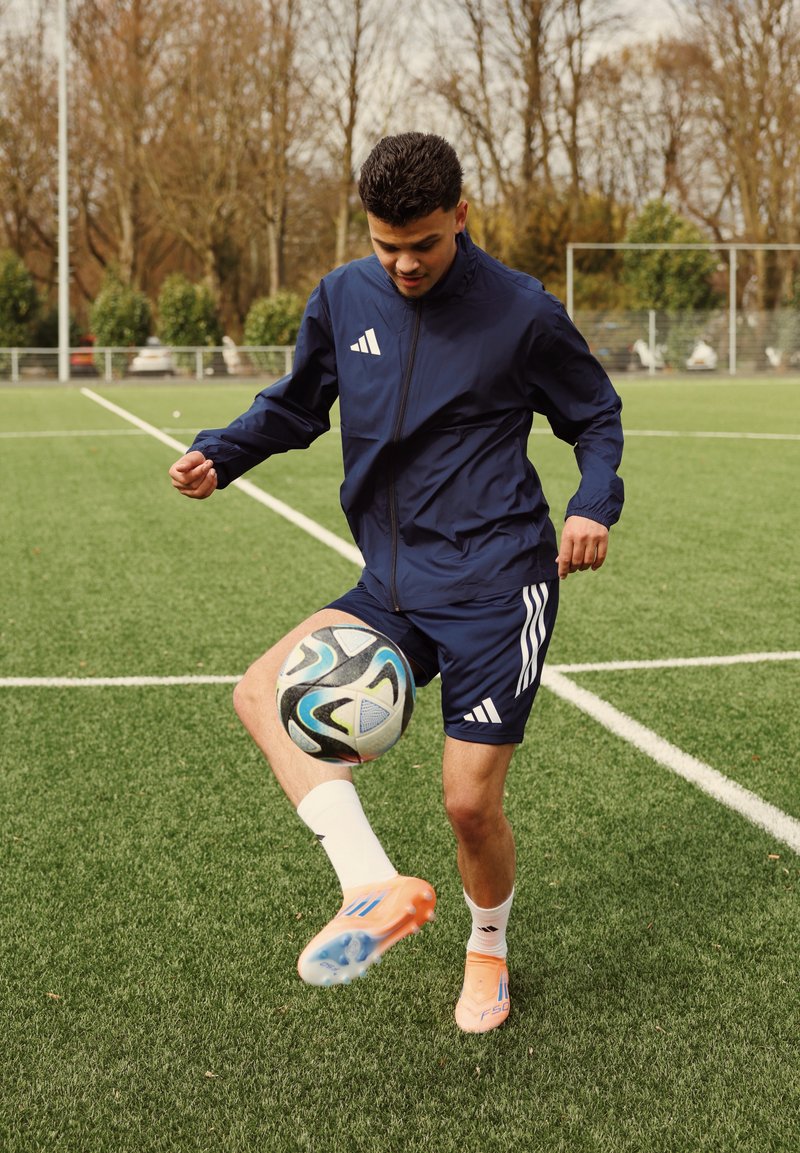 Jeune homme en tenue de sport bleu marine jonglant avec un ballon de football sur un terrain en gazon vert, avec des arbres dépourvus de feuilles et des poteaux de but en arrière-plan.