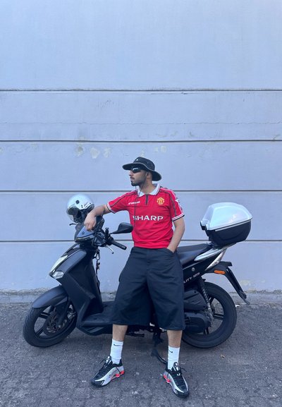 Scooter negra con un casco plateado aparcado contra una pared gris. Persona con una camiseta roja y pantalones cortos negros se apoya de manera casual contra la scooter.