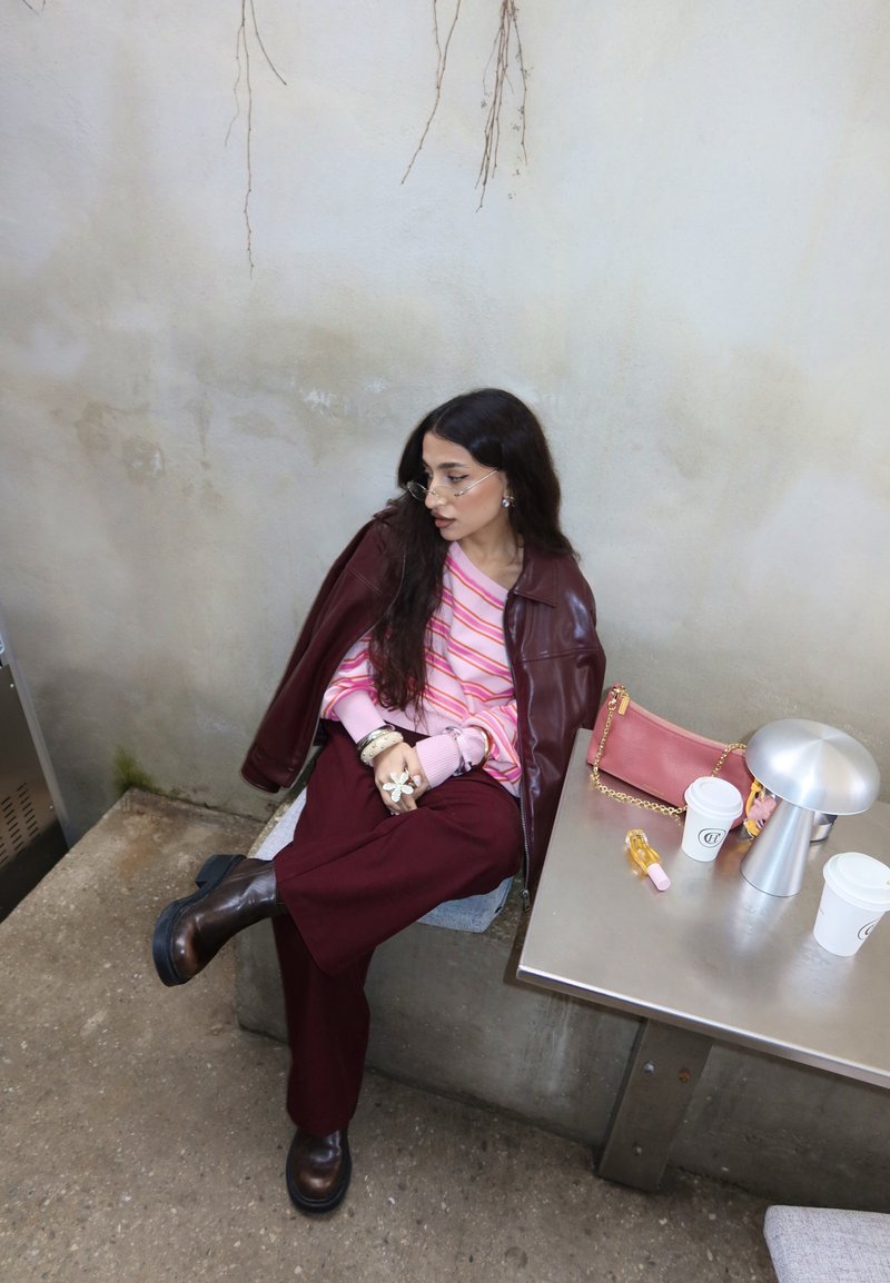 Femme en pull rayé rose et pantalon bordeaux assise jambes croisées à côté d'une table tenant un sac à main rose, des tasses de café et une petite lampe.