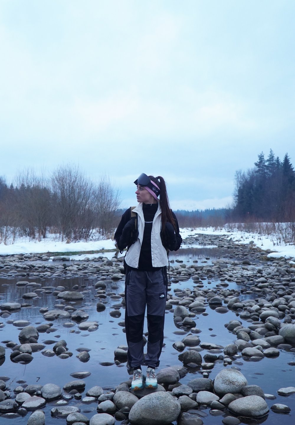 Fille debout sur des rochers près d'un ruisseau, portant un gilet beige sur une chemise noire, un pantalon gris et des chaussures bleu clair ; arrière-plan nuageux.