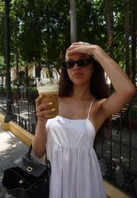 White dress with thin straps, holding an iced coffee with a green straw. Black sunglasses, yellow nail polish, and a black handbag.