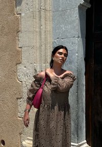 Καφέ off-shoulder φόρεμα με πρησμένα μακριά μανίκια που διαθέτει σχέδιο eyelet, συνδυασμένο με ροζ τσάντα και ασημένιο βραχιόλι, μπροστά σε τοίχο από πέτρα.