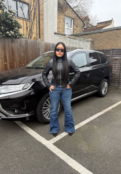 Chaqueta de cuero negra con cremallera, jeans de campana azul y tacones de punta negros. SUV negro aparcado de fondo, cerca de madera, pavimento gris.