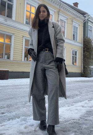 Mujer con abrigo largo gris, suéter de cuello alto negro, pantalones grises, guantes negros y botas, de pie en una calle nevada con un edificio amarillo de madera detrás.