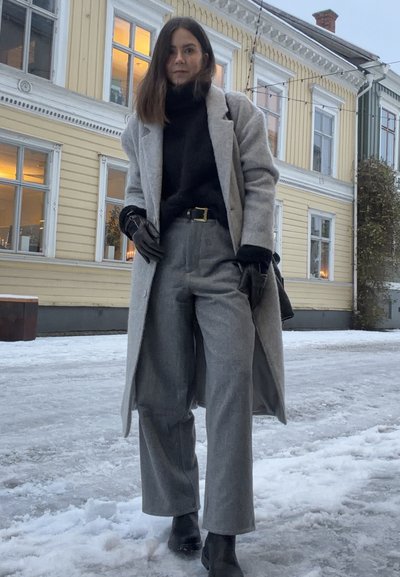 Mujer con abrigo largo gris, suéter de cuello alto negro, pantalones grises, guantes negros y botas, de pie en una calle nevada con un edificio amarillo de madera detrás.