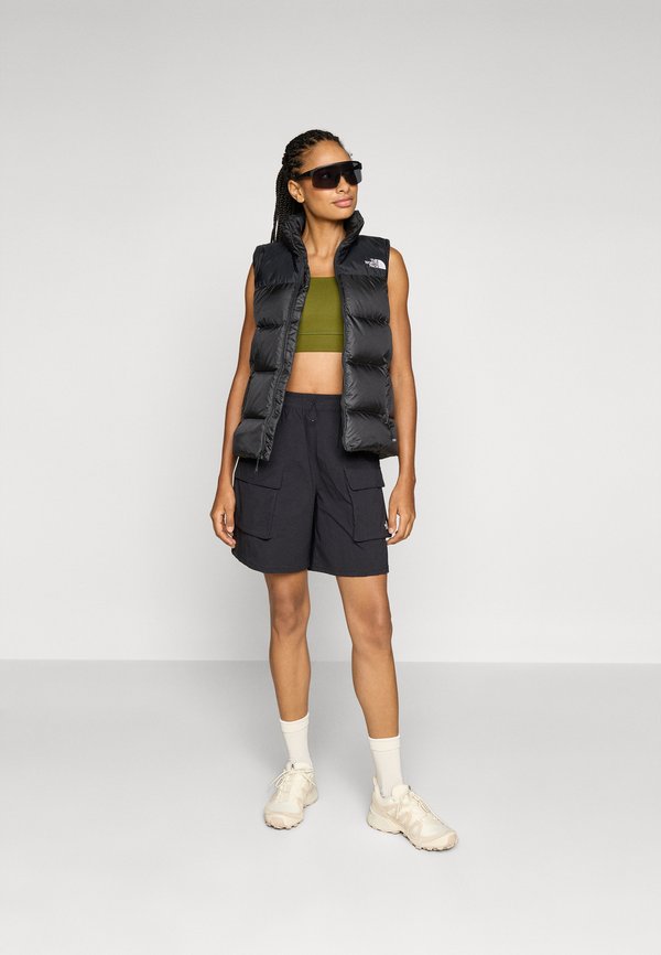 Crna pufasta prsluk s visokim ovratnikom, maslinasto zeleni crop top i crne cargo šortse. Bijele tenisice i sunčane naočale upotpunjuju outfit.
