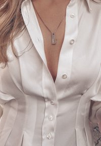 Chemise en satin blanc avec une rangée de boutons, présentant un col en V. Un collier argenté avec un pendentif rectangulaire pend au niveau du décolleté.