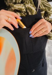 Mains avec des ongles bleus à motifs rangeant un baume à lèvres orange dans une poche zippée noire d'une veste, personne portant une polaire verte et tenant un snowboard.