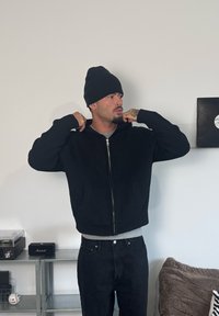 Melna adīta rāvējslēdzēja jaka ar kapuci, nēsāta pār pelēku kreklu. Dizainā ir riboti aproču gali un kabatas, kopā ar melnām biksēm.