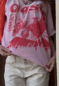 Άτομο που φοράει ροζ μπλουζάκι με το λογότυπο του συγκροτήματος Oasis, με κόκκινα γραφικά που αναφέρονται στο άλμπουμ «Definitely Maybe» και παντελόνι κρεμ χρώματος.