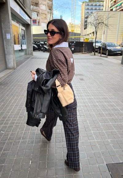 Mujer con conjunto de cuadros y gafas de sol camina por la acera de la ciudad llevando una chaqueta de cuero negra y un bolso beige colgados del hombro.