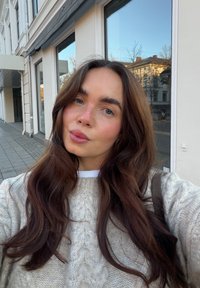 Nærbillede af en person med langt, bølget brunt hår iført en lysegrå kabelstrikket sweater over en hvid skjorte. Baggrunden viser byens bygninger.