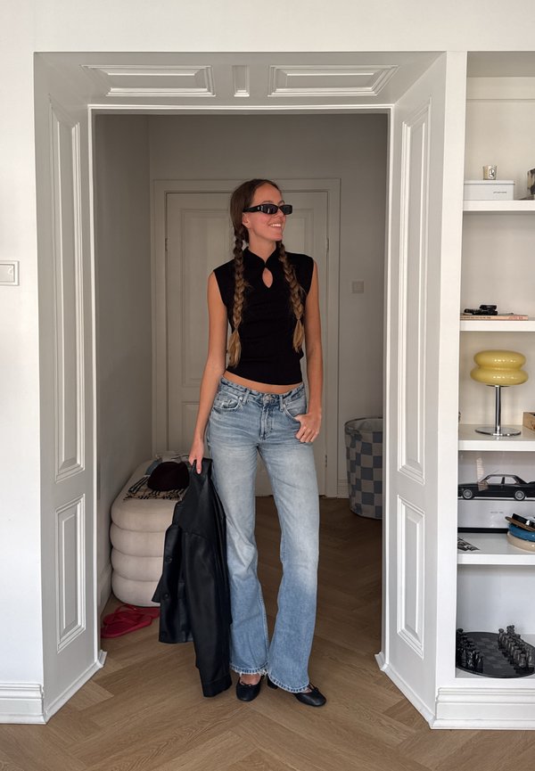 Mujer con trenzas largas, gafas de sol, camiseta negra sin mangas, vaqueros azul claro y sosteniendo una chaqueta negra, de pie en un umbral en el interior.
