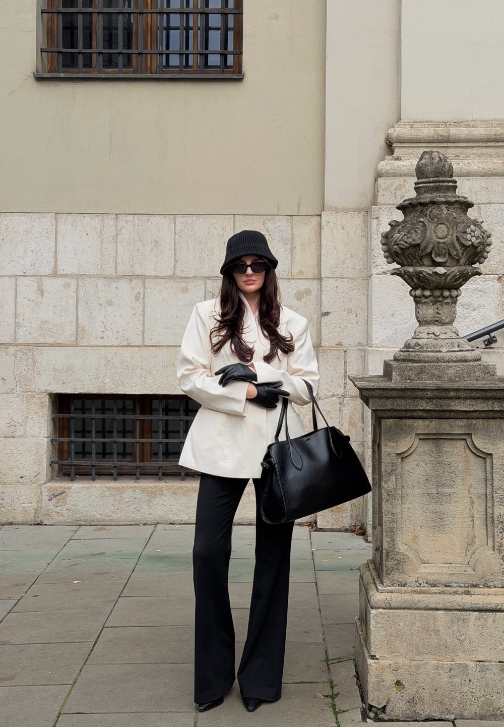 Blazer beige clair sur un haut noir, pantalon noir évasé, chapeau seau noir et gants noirs. Tient un sac à main noir structuré.