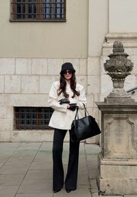 Blazer beige clair sur un haut noir, pantalon noir évasé, chapeau seau noir et gants noirs. Tient un sac à main noir structuré.