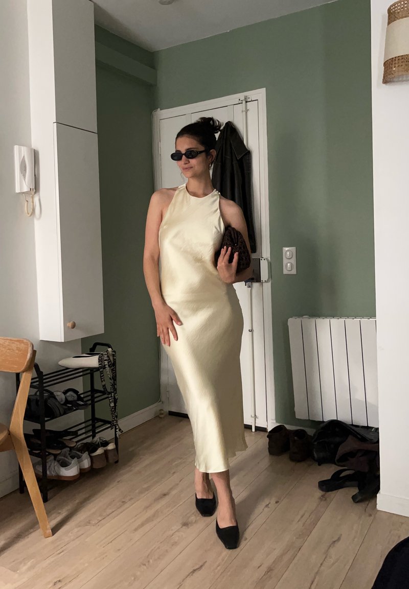 Femme portant une robe en satin crème sans manches, des lunettes de soleil noires et des talons, tenant une pochette, debout dans une pièce avec des murs verts et un sol en bois.