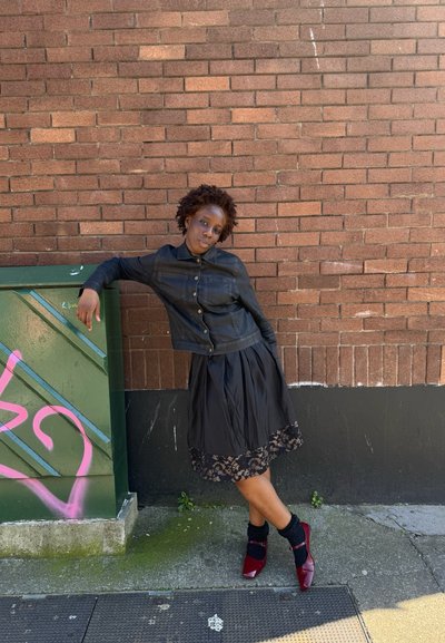 Mujer con chaqueta negra y falda con borde de encaje apoyada en una caja de servicios pública verde cubierta de grafitis contra una pared de ladrillos, lleva zapatos rojos y calcetines negros.