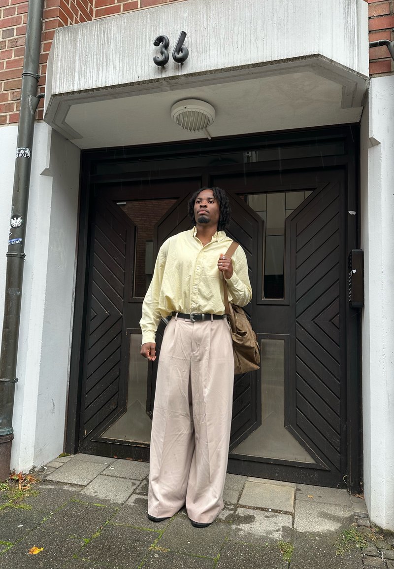 Un homme avec des dreadlocks portant une chemise jaune ample et un pantalon large beige se tient devant des portes noires à double battant sous un immeuble portant le numéro 36.