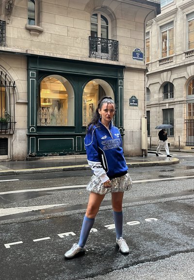 Chaqueta azul con cremallera y acentos blancos, falda plateada, calcetas altas azules y zapatos plateados. Entorno urbano con un fondo de edificio.