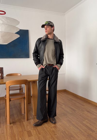 Joven con camisa a rayas, chaqueta de cuero negra con cuello de piel, pantalones anchos negros y gorra, de pie en un comedor minimalista con suelo de madera.
