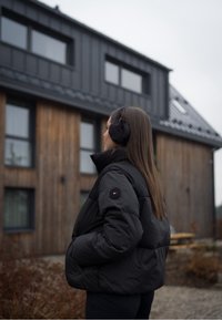 Casaco puffer preto com colarinho alto e costuras visíveis, combinado com auriculares de pele sintética pretos. Ambiente exterior com fundo de madeira.