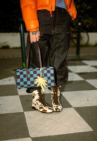 Personne portant un pantalon noir, une veste orange, des bottes à motif vache, tenant un sac à carreaux bleu et marron avec un pompon jaune, debout sur un sol carrelé.