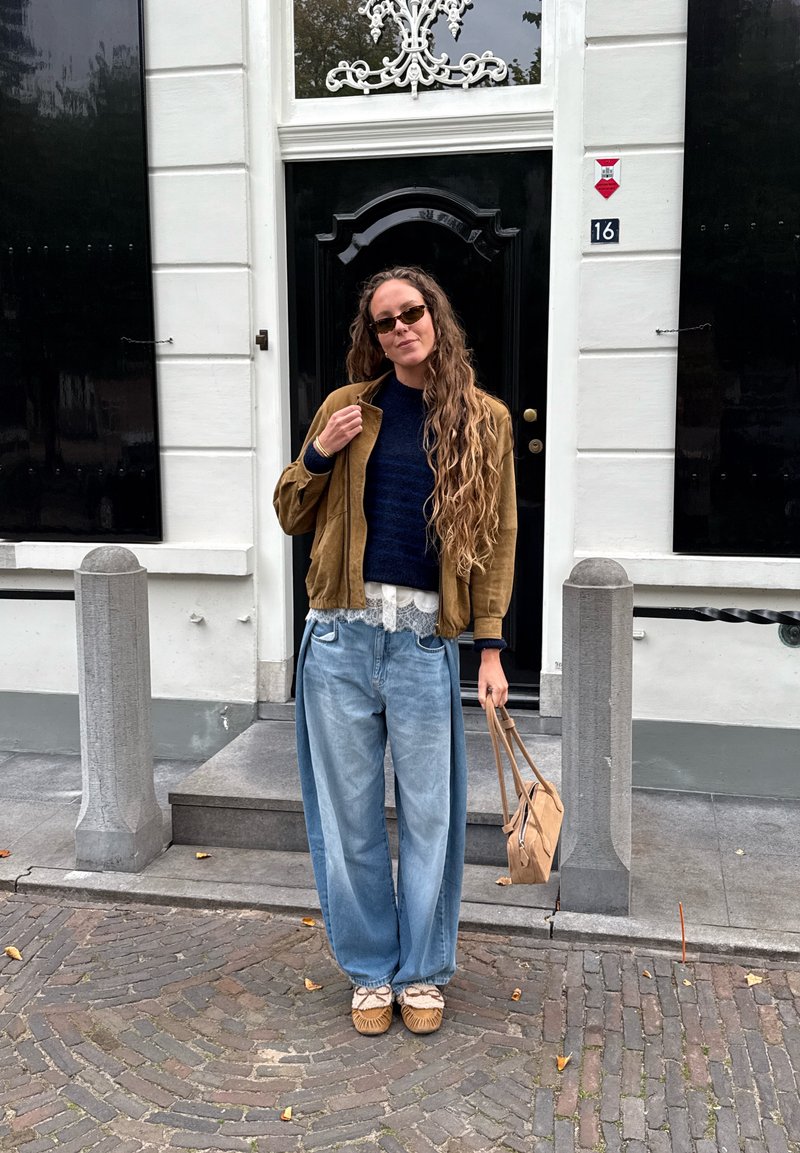 Giacca bomber in suede marrone, maglione lavorato a maglia blu navy, rifinitura in pizzo bianco, jeans oversize in denim azzurro chiaro, borsa marrone chiaro e calzature color cuoio.