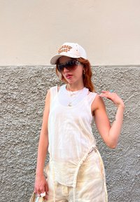 Λευκή αμάνικη μπλούζα με συγκεντρωμένη λεπτομέρεια στο πλάι, ανοιχτό μπεζ παντελόνι, λευκό καπέλο με πορτοκαλί κείμενο, σκούρα γυαλιά ηλίου, στρωματοποιημένα κολιέ.