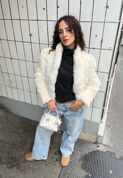 Chaqueta de piel sintética blanca, suéter de cuello alto negro, vaqueros holgados azul claro, botines color canela, bolso blanco pequeño con herrajes, fondo de azulejos texturizados.