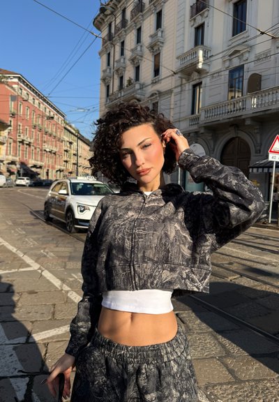 Mujer con cabello rizado que lleva una sudadera corta con capucha negra y pantalones con estampado, de pie en una calle soleada de la ciudad con edificios históricos y coches.