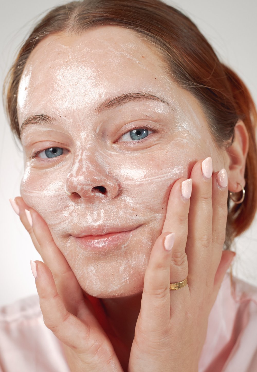 Zbliżenie na twarz z lekką, pienistą maską na twarz. Gładka, promienna skóra i delikatne różowe paznokcie, z rękami delikatnie dotykającymi twarzy.